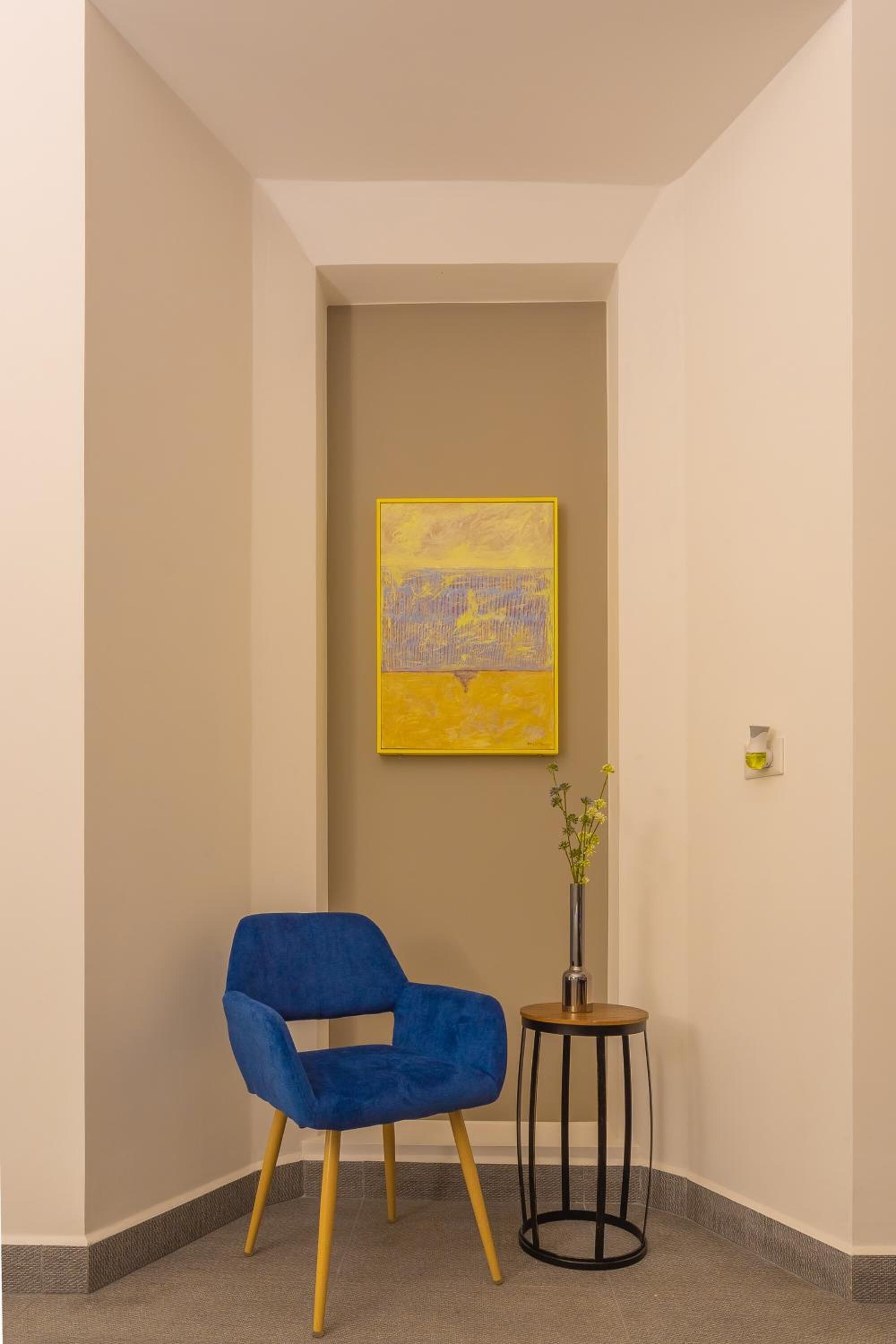 Um canto de um quarto com uma cadeira azul, uma pequena mesa redonda com um vaso e um quadro amarelo na parede.