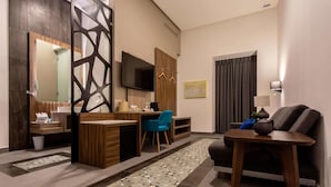 Une chambre d’hôtel moderne avec un bureau en bois, un téléviseur à écran plat, une chaise bleue, un canapé et un panneau décoratif mural.