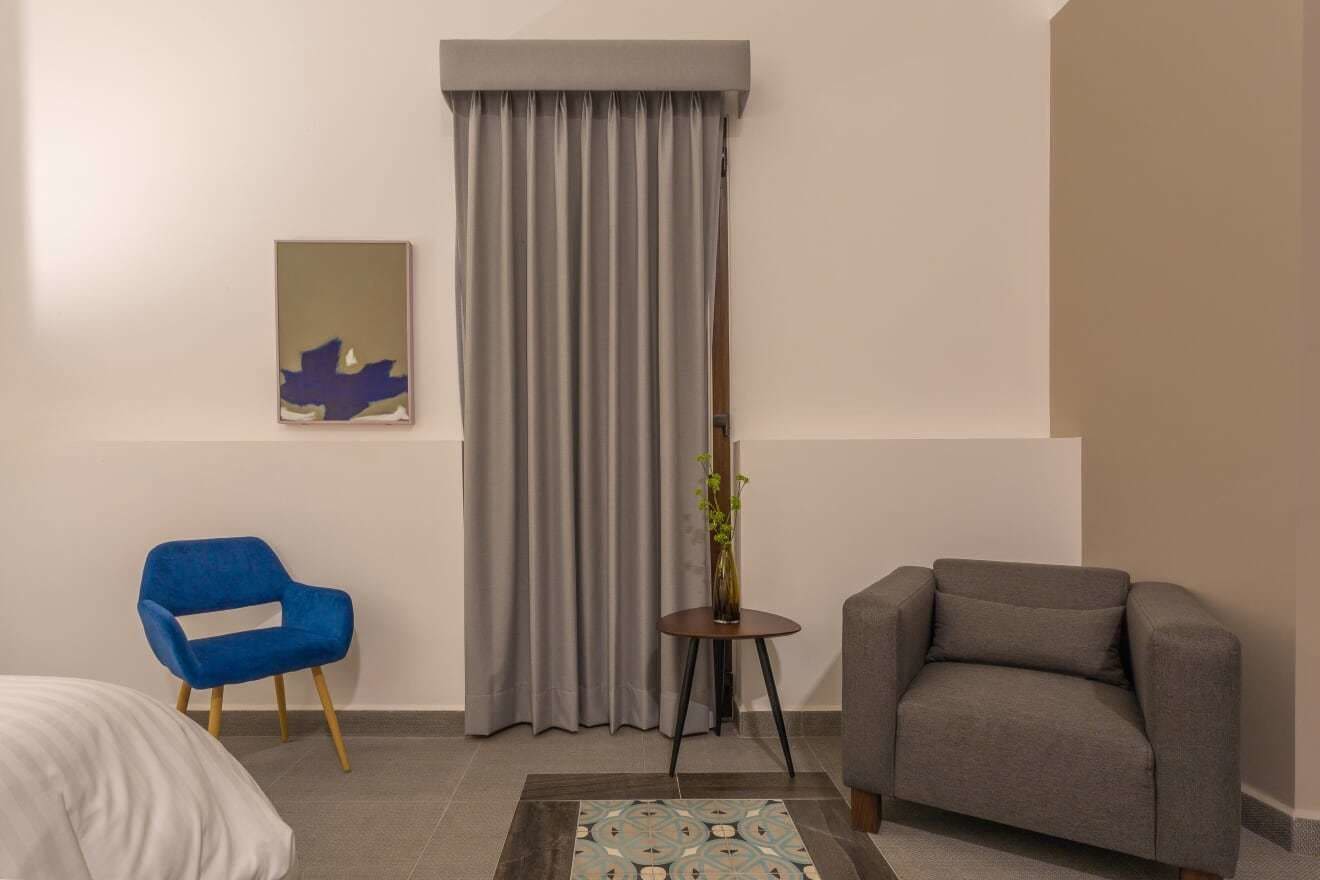Une chambre d’hôtel avec un lit, une chaise bleue, un fauteuil gris, un rideau gris, une petite table avec un vase et un tableau encadré au mur.