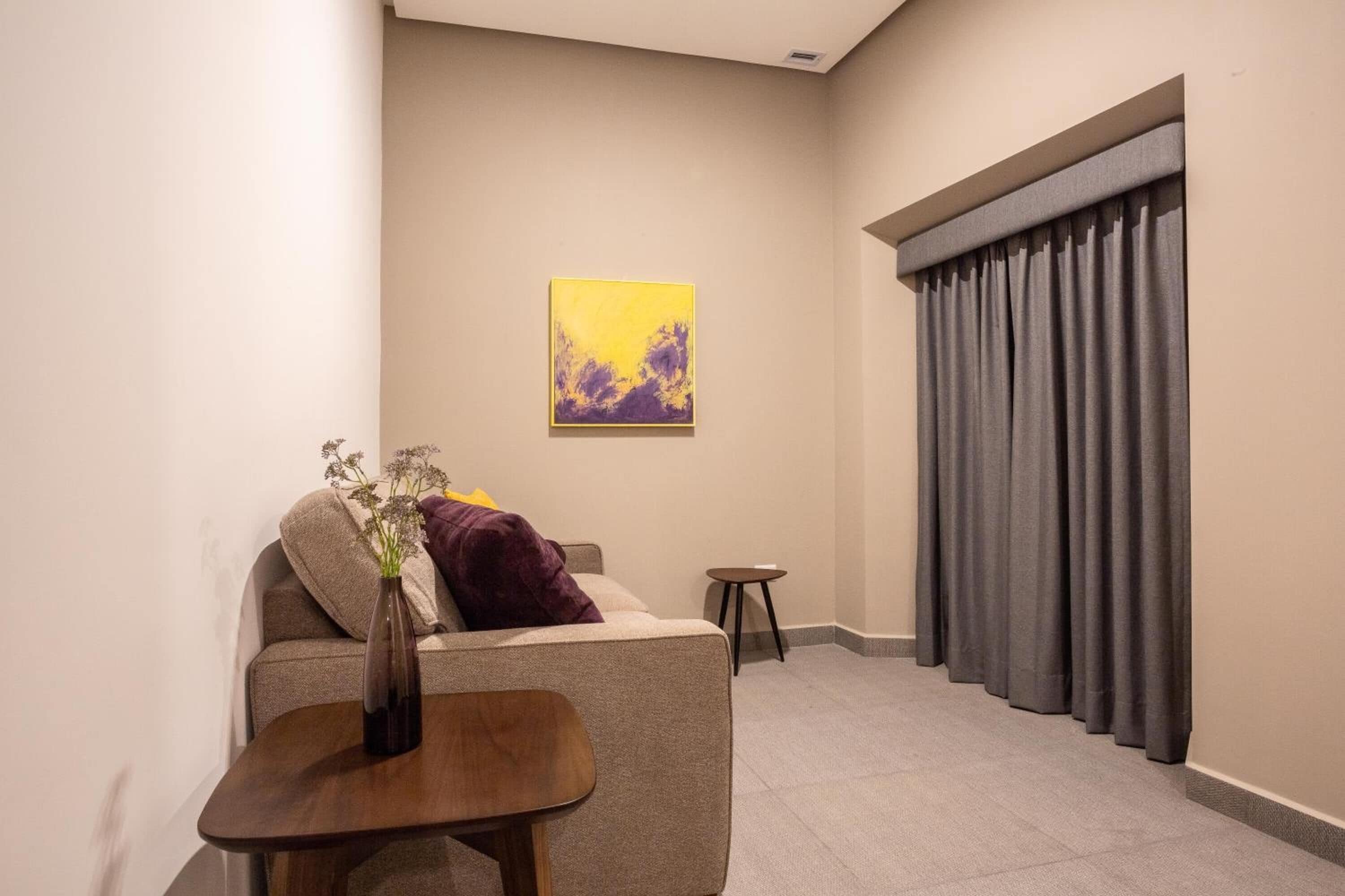 Una sala moderna con un sofá, una mesita, un jarrón con flores, un cuadro en la pared y un ventanal con cortinas.