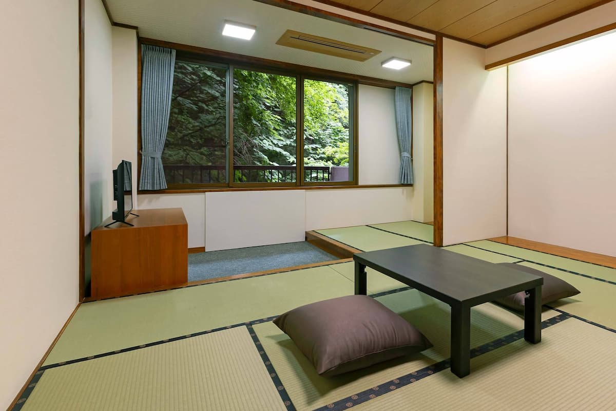 Une pièce avec un sol recouvert de tatamis, une petite table basse, des coussins, un bureau en bois et une grande fenêtre avec des rideaux.