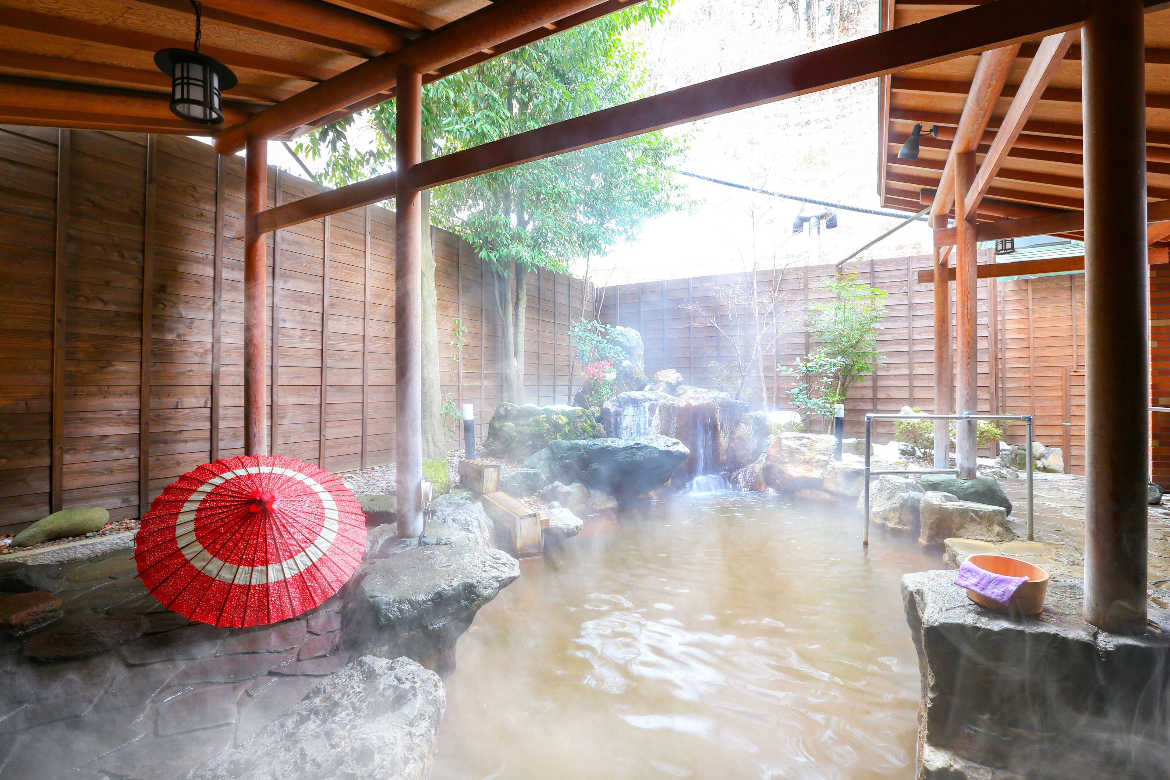 Ein traditionelles japanisches Onsen mit Holzinnenraum, einem Steine-Thermalbad und einem roten Sonnenschirm.