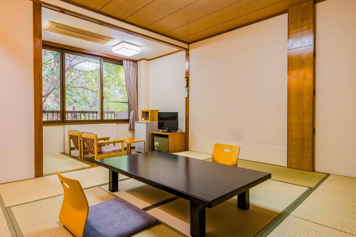 A traditional Japanese room with tatami flooring, a low table, chairs, a television, and a window with a view of trees.