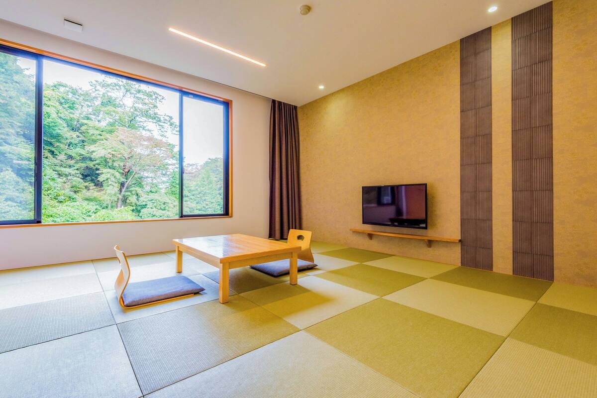 A modern room with tatami flooring, a wooden table, a TV on a shelf, and a large window with a view of greenery.