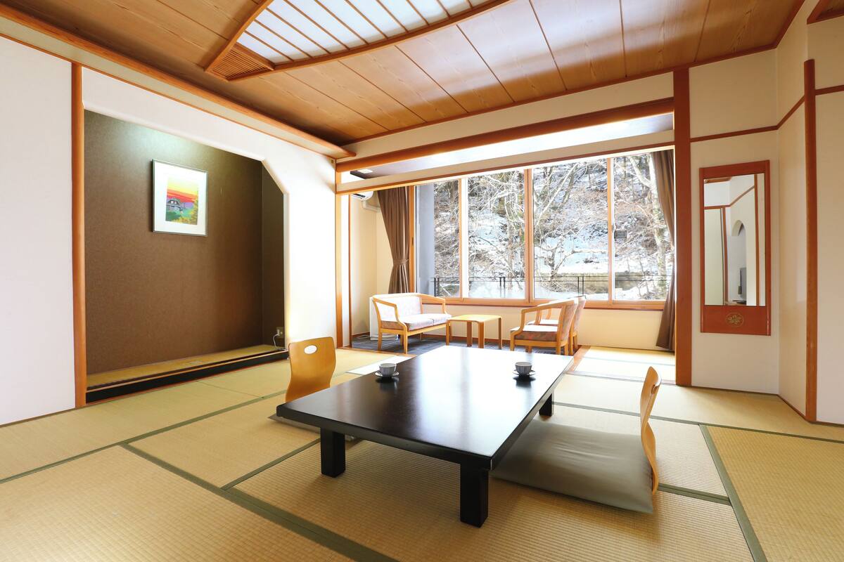 A traditional Japanese room with tatami flooring, a low table, and a sliding door leading to a balcony with a view of trees.