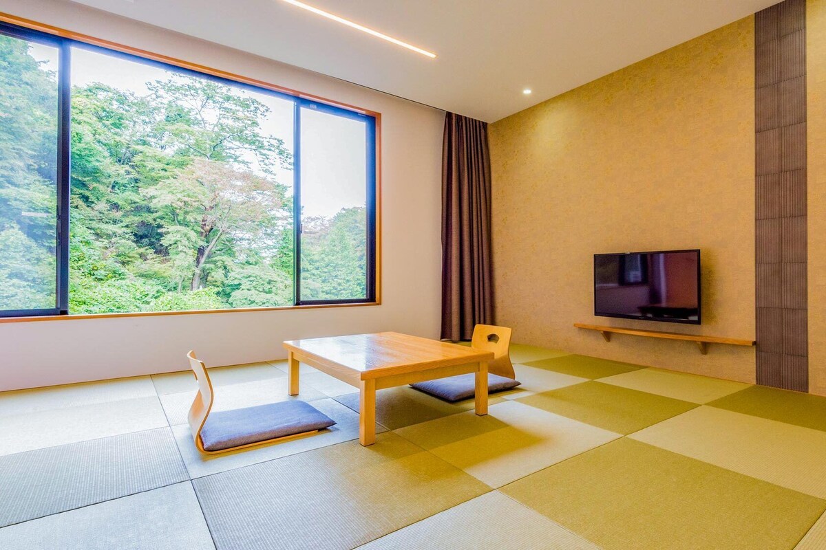 A room with tatami mats, a low table, a chair, a TV on a shelf, and a large window with a view of trees.