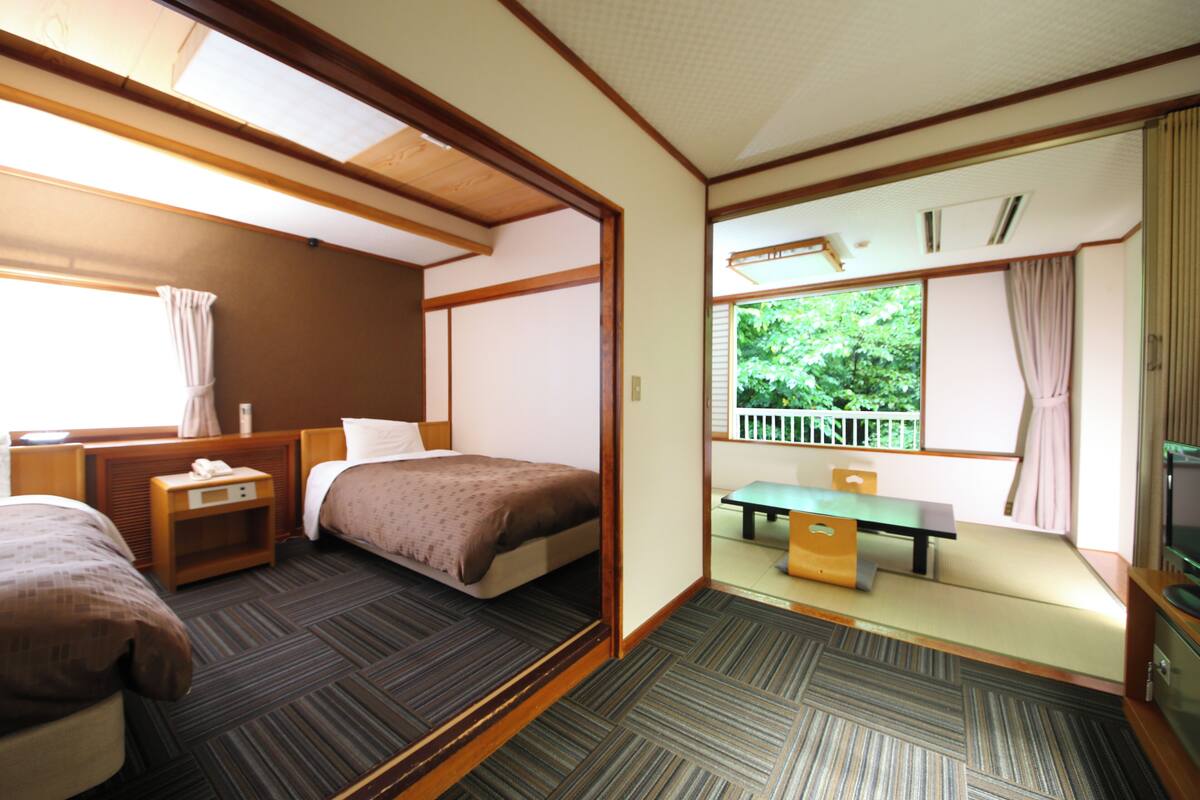 A hotel room with two beds, a small table, a bench, and a window with a view of greenery.