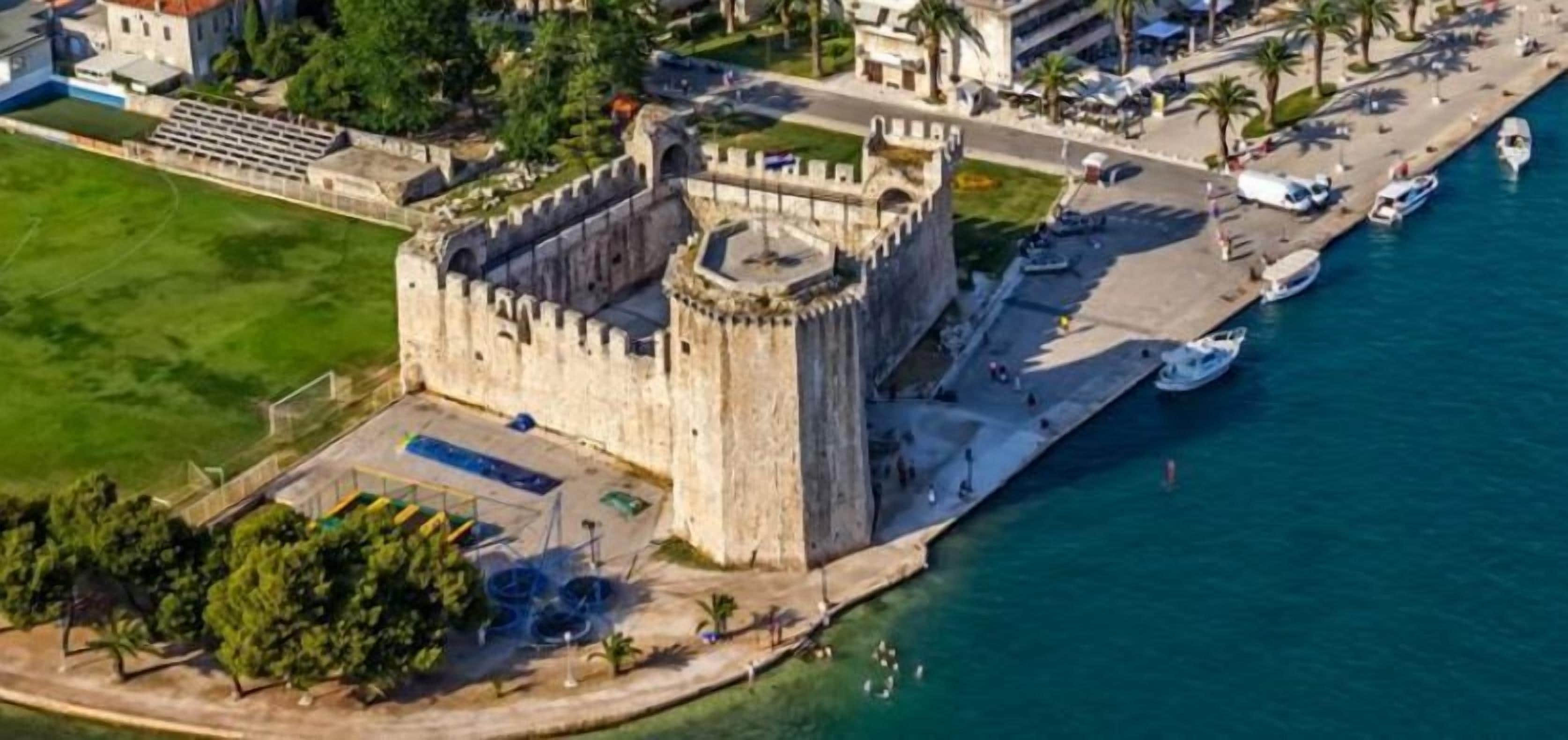 An aerial view of a historic castle with a fortified structure, a waterfront promenade, and a small harbor with boats.