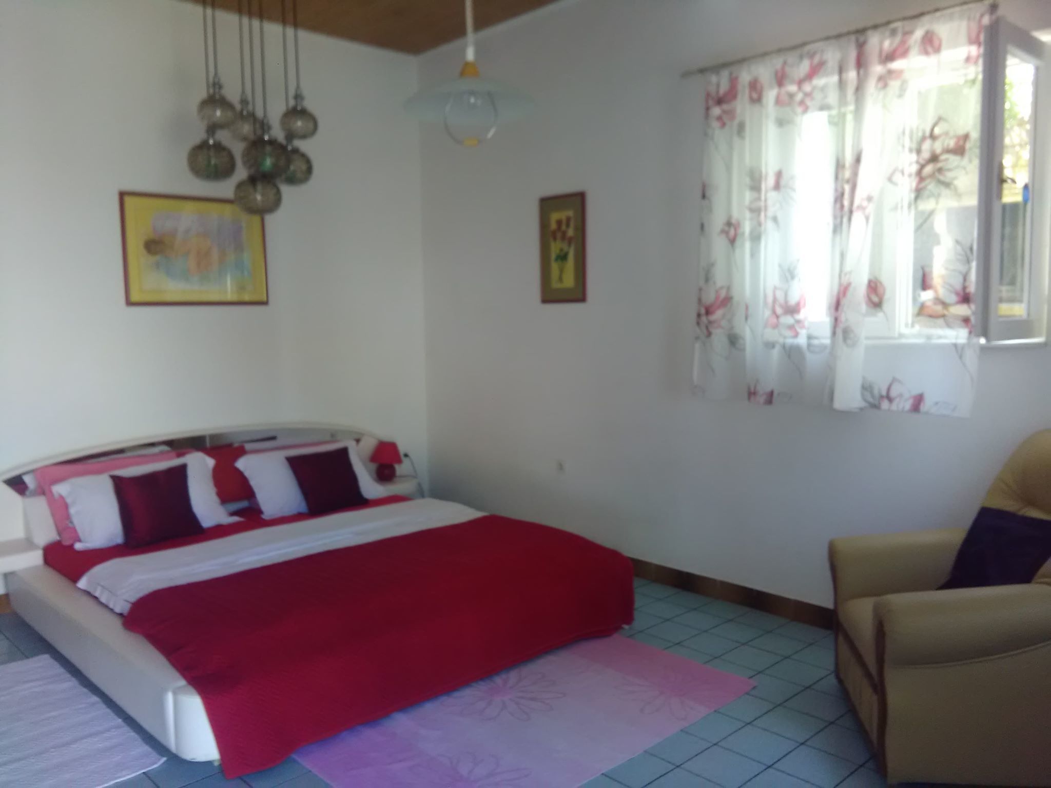 Un dormitorio con cama, una silla, ventana con cortinas florales y lámpara colgante.