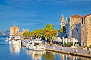Una marina frente al agua con barcos amarrados, un edificio de piedra con una torre y palmeras bordeando el paseo.