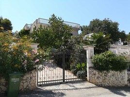 Una entrada protegida con muro de piedra y portón metálico, rodeada de abundante vegetación y un edificio al fondo.