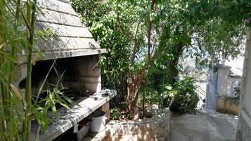 A stone barbecue grill with a stone bench, surrounded by greenery and flowering plants.