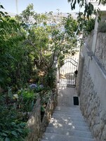 Una escalera de piedra que conduce a una entrada con puerta, rodeada de abundante vegetación y vista al mar.