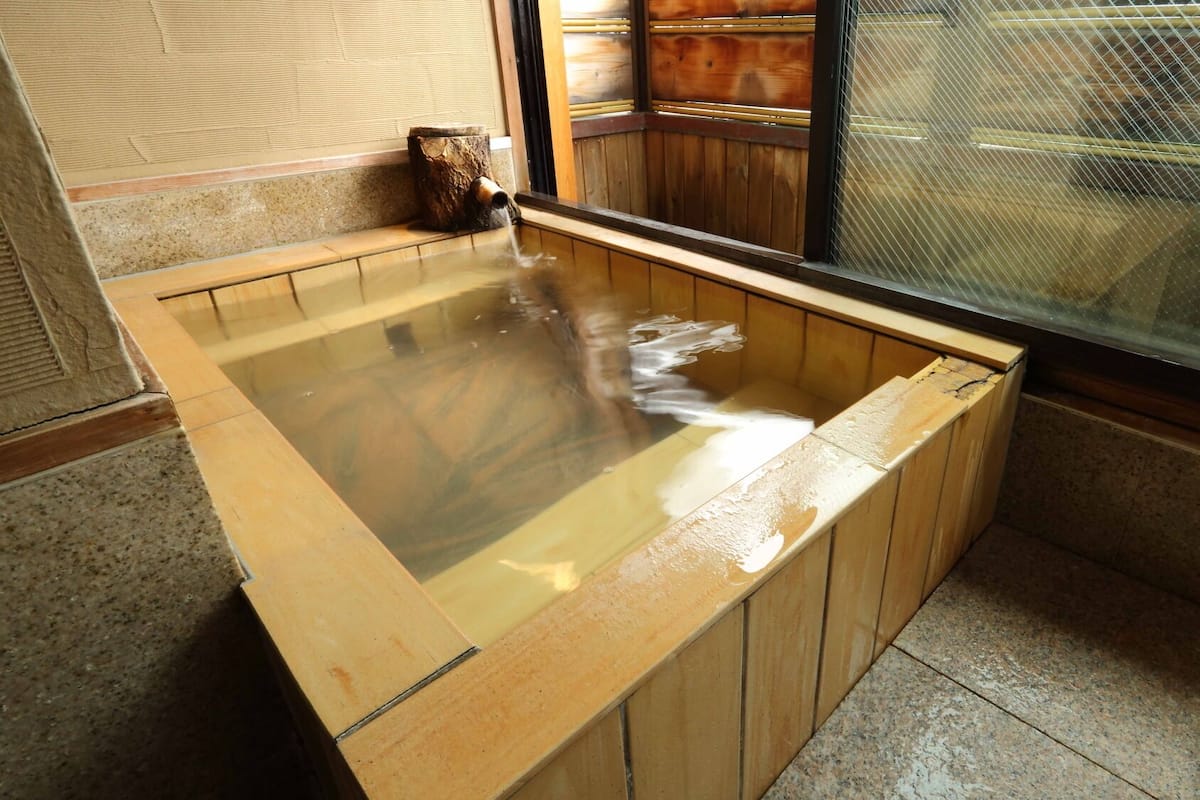 A wooden hot tub with a wooden bench, a small wooden stool, and a window with a metal screen.