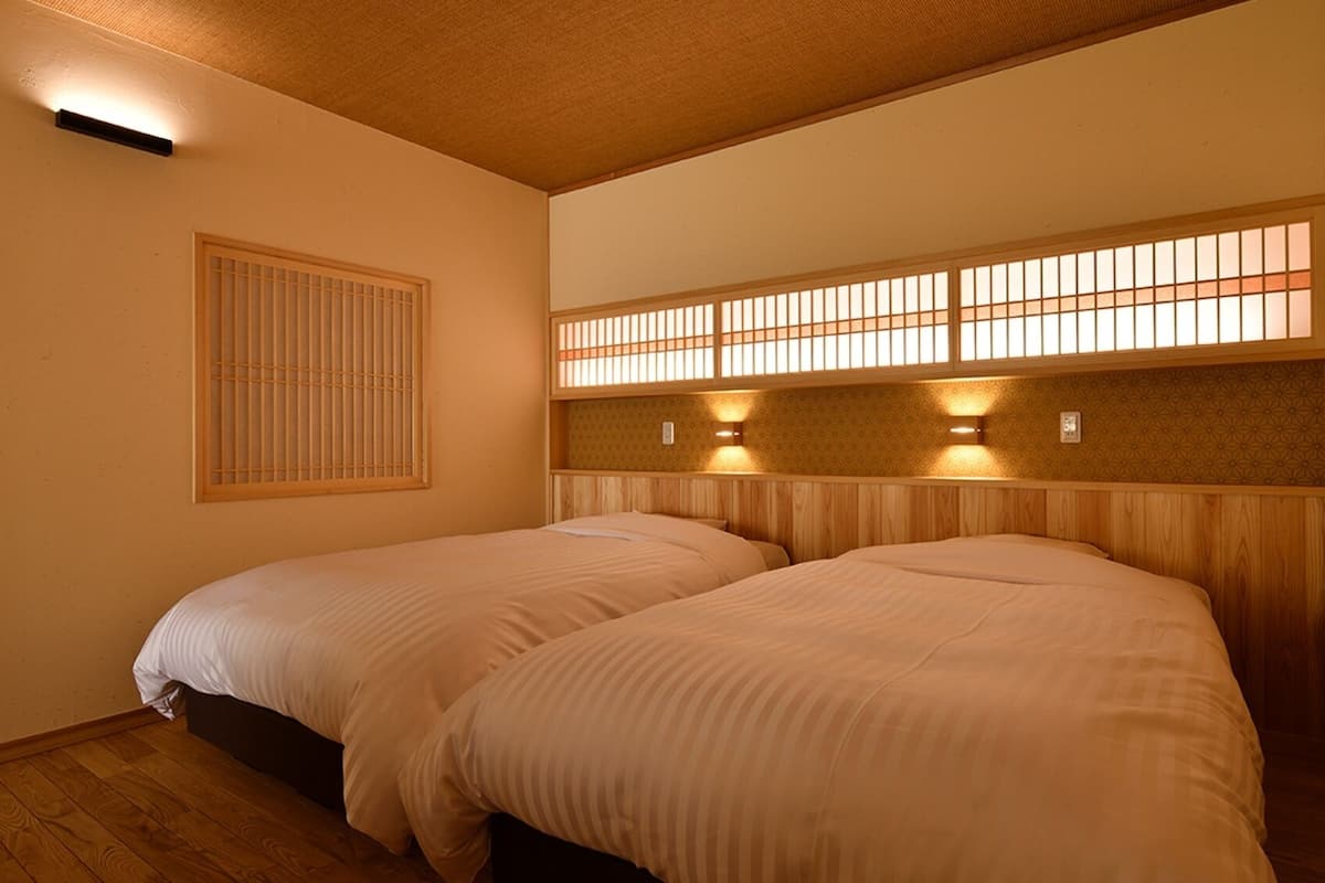 A room with two beds, a window with a wooden frame, and a wall-mounted light fixture.