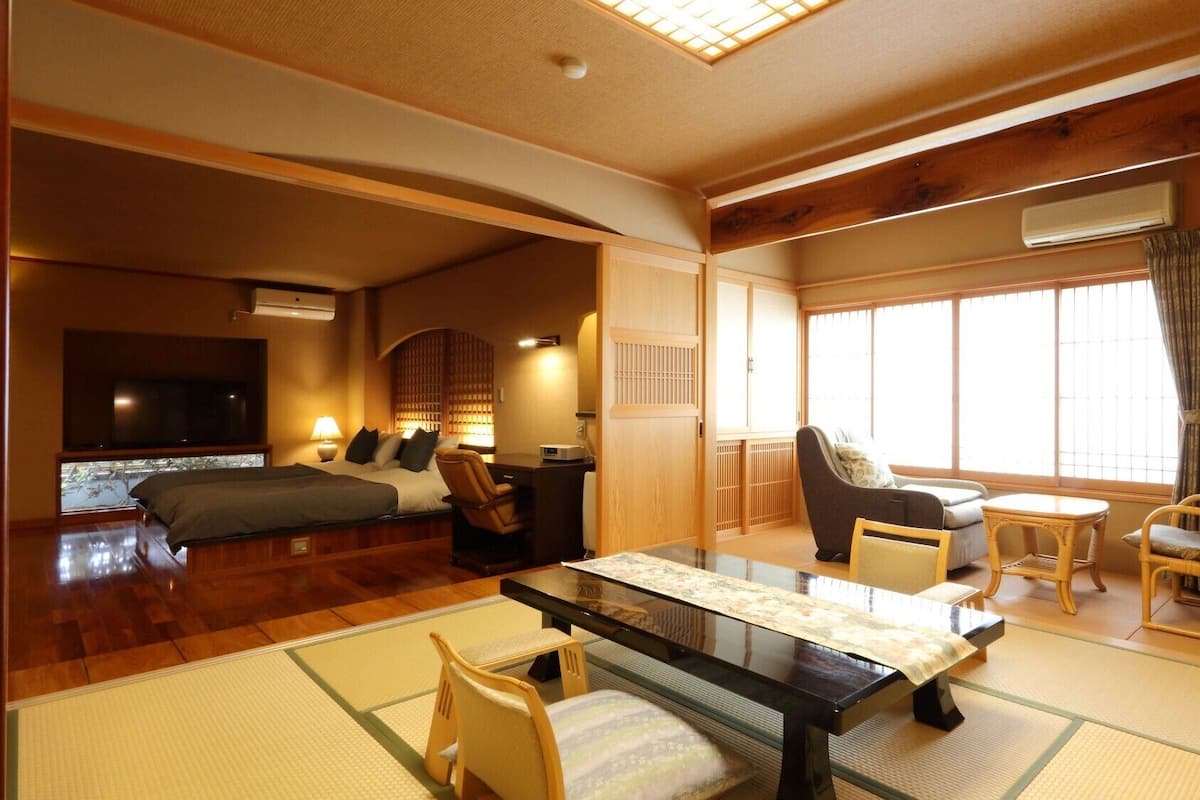 A traditional Japanese-style room with tatami flooring, a low table, chairs, a bed, and a television.