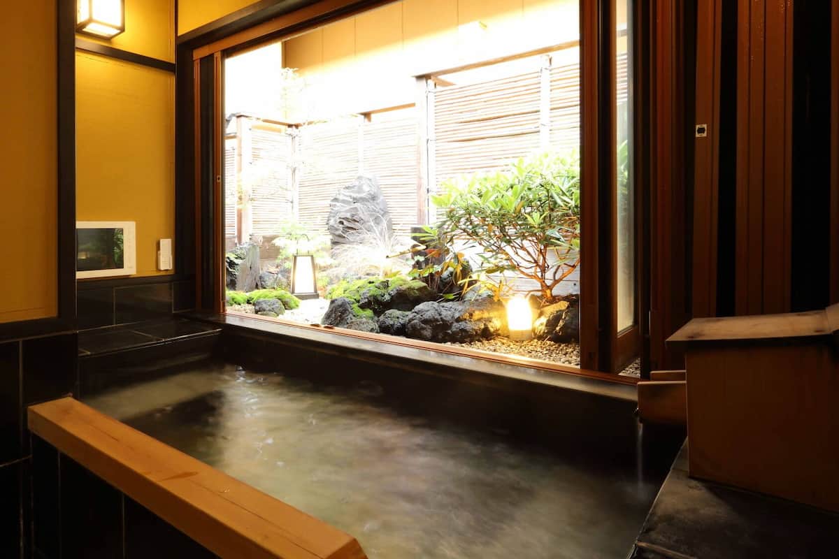 A traditional Japanese-style bathroom with a stone basin, wooden bench, and a view of a garden through a window.