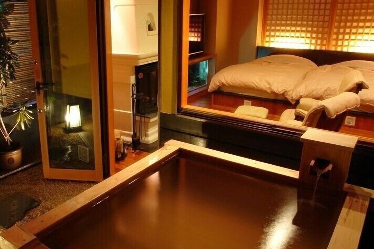 A traditional Japanese onsen with a wooden bath, a sliding glass door, and a bed with white bedding.