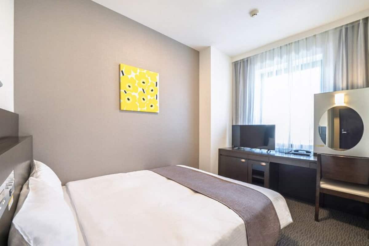 A hotel room with a bed, a desk, a chair, a TV, a mirror, and a window with curtains.