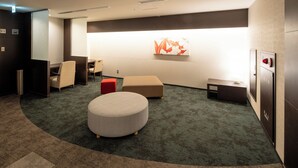 A hotel lobby with a curved carpet, a round ottoman, a red stool, a desk, and a wall-mounted artwork. - Osaka Airterminal Hotel (Toyonaka)