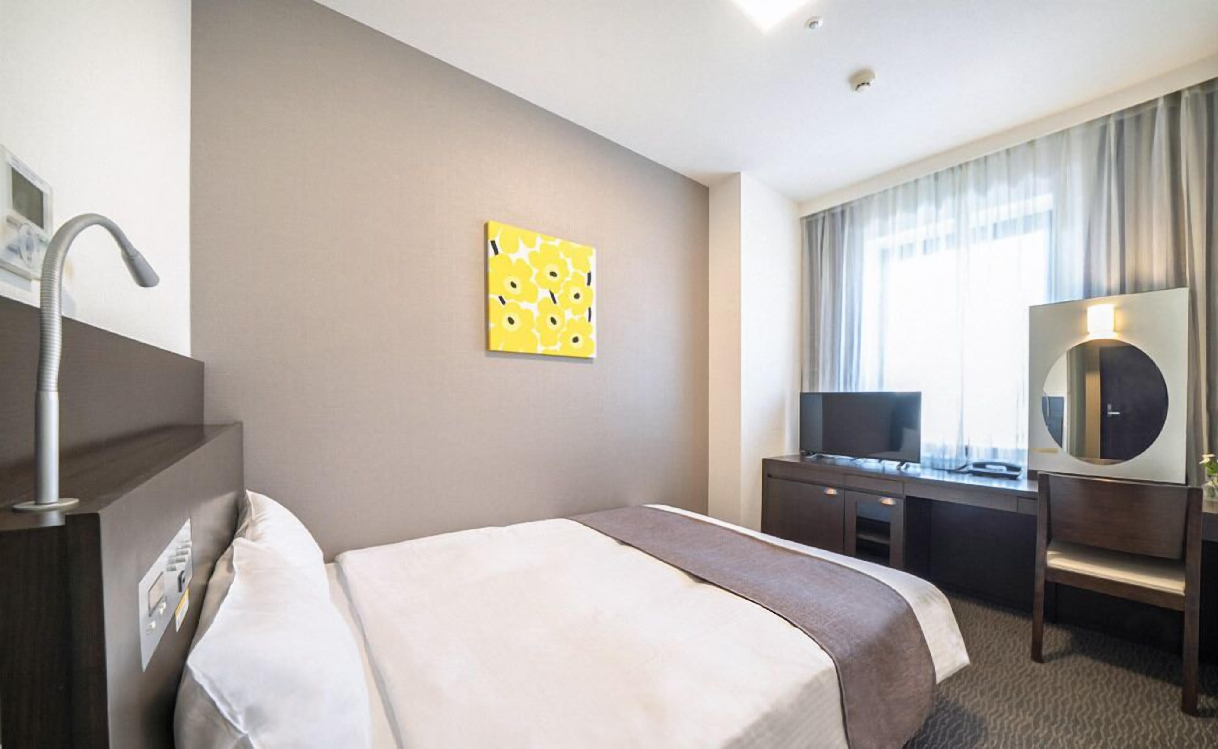 A hotel room with a bed, a desk with a TV, a mirror, and a window with curtains.