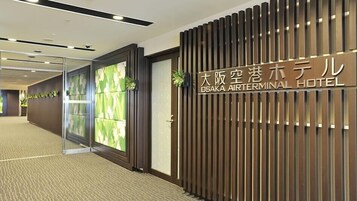 The interior of the Osaka Airterminal Hotel with a reception area, a wooden wall with the hotel's name, and a glass door with a view of the interior.