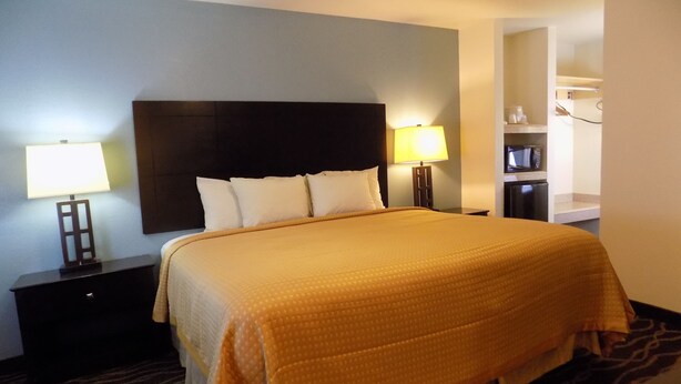 A hotel room with a large bed, two bedside lamps, a dark headboard, and a small kitchenette in the corner. - Budget Inn (Tucumcari)