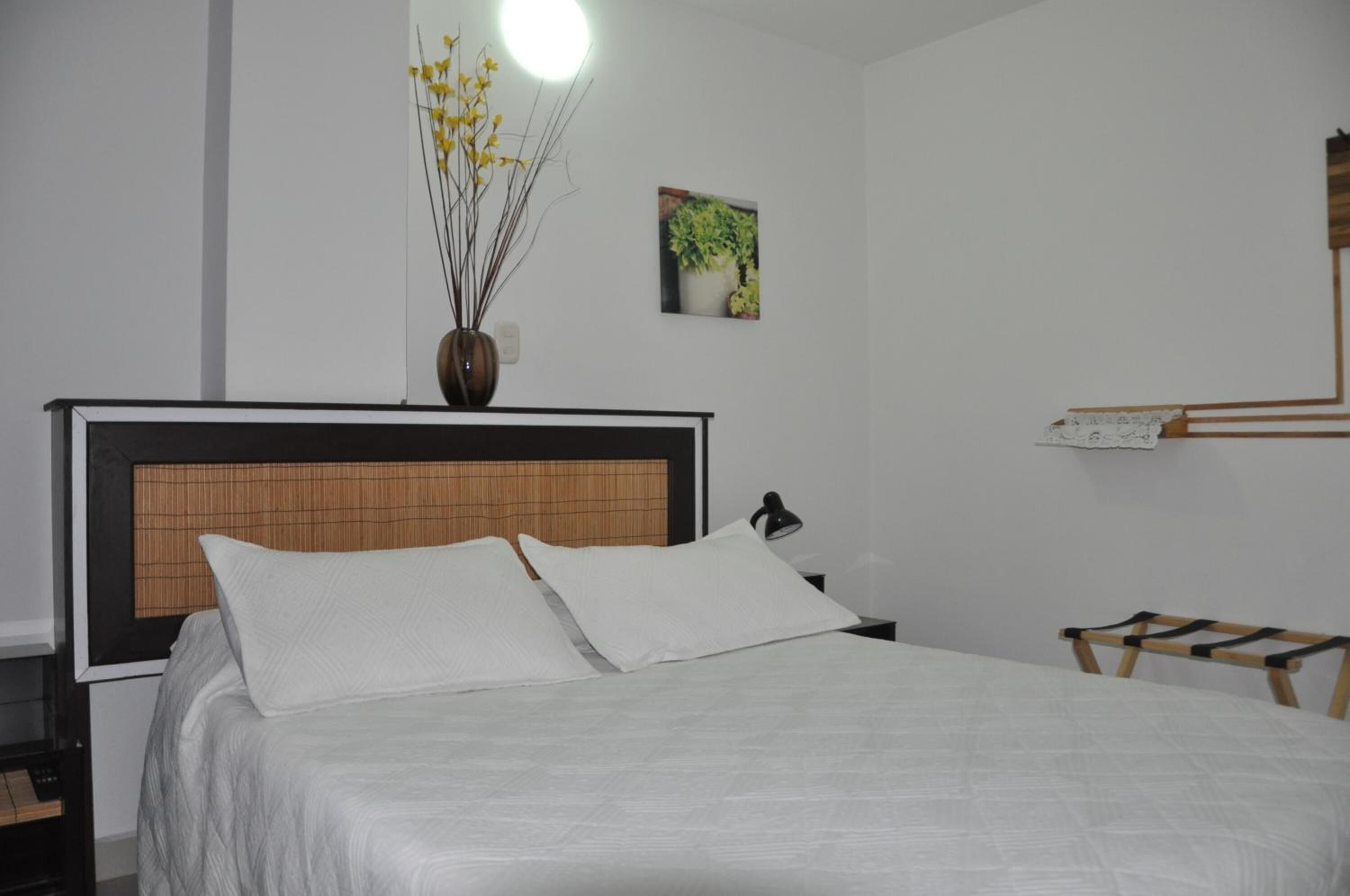 A neatly made bed with white linens, a bedside lamp, a vase with yellow flowers, and a small potted plant on the wall.