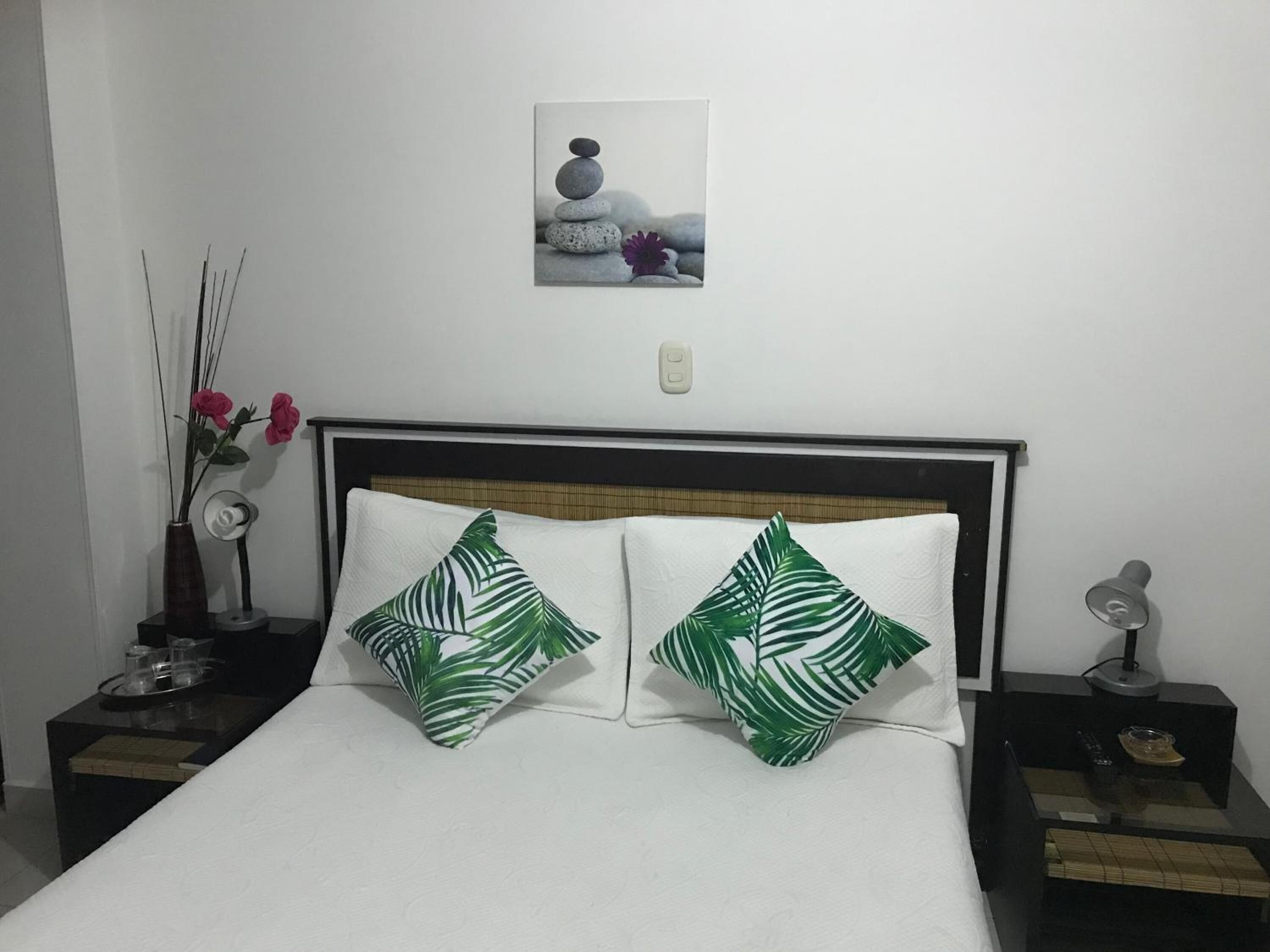 A neatly made bed with two green leaf-patterned pillows, a bedside table with a vase of flowers, and a wall-mounted picture of stones and flowers.