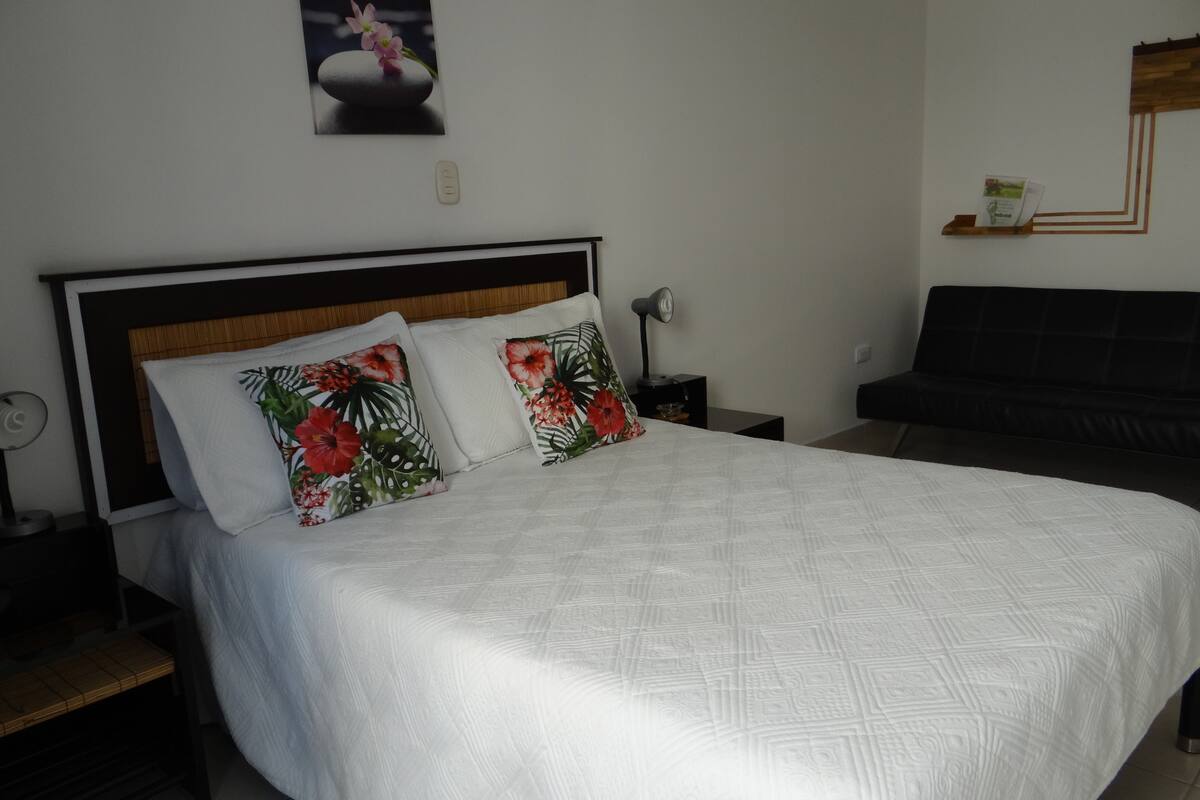 Una cama bien tendida con colcha blanca y dos almohadas florales, una mesita de noche con lámpara y un sofá oscuro contra una pared blanca.