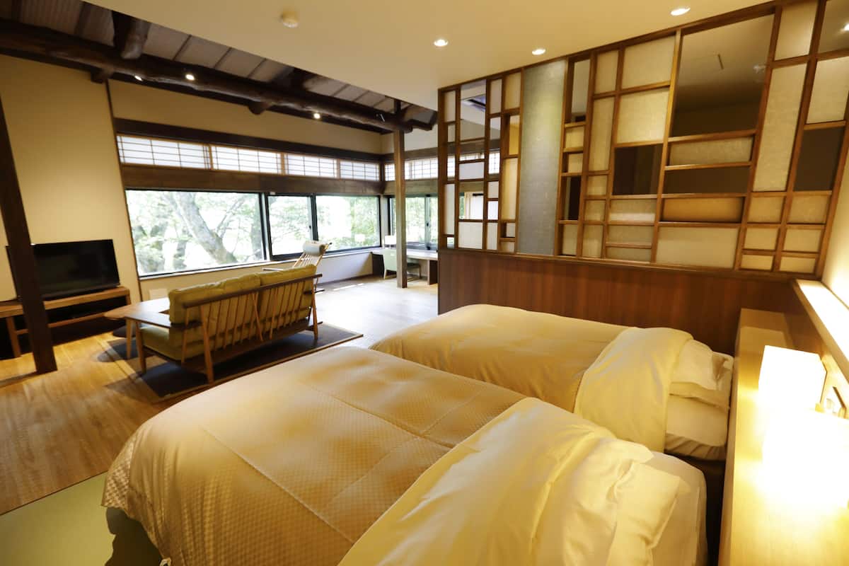 A hotel room with two beds, a TV, a desk, and large windows.