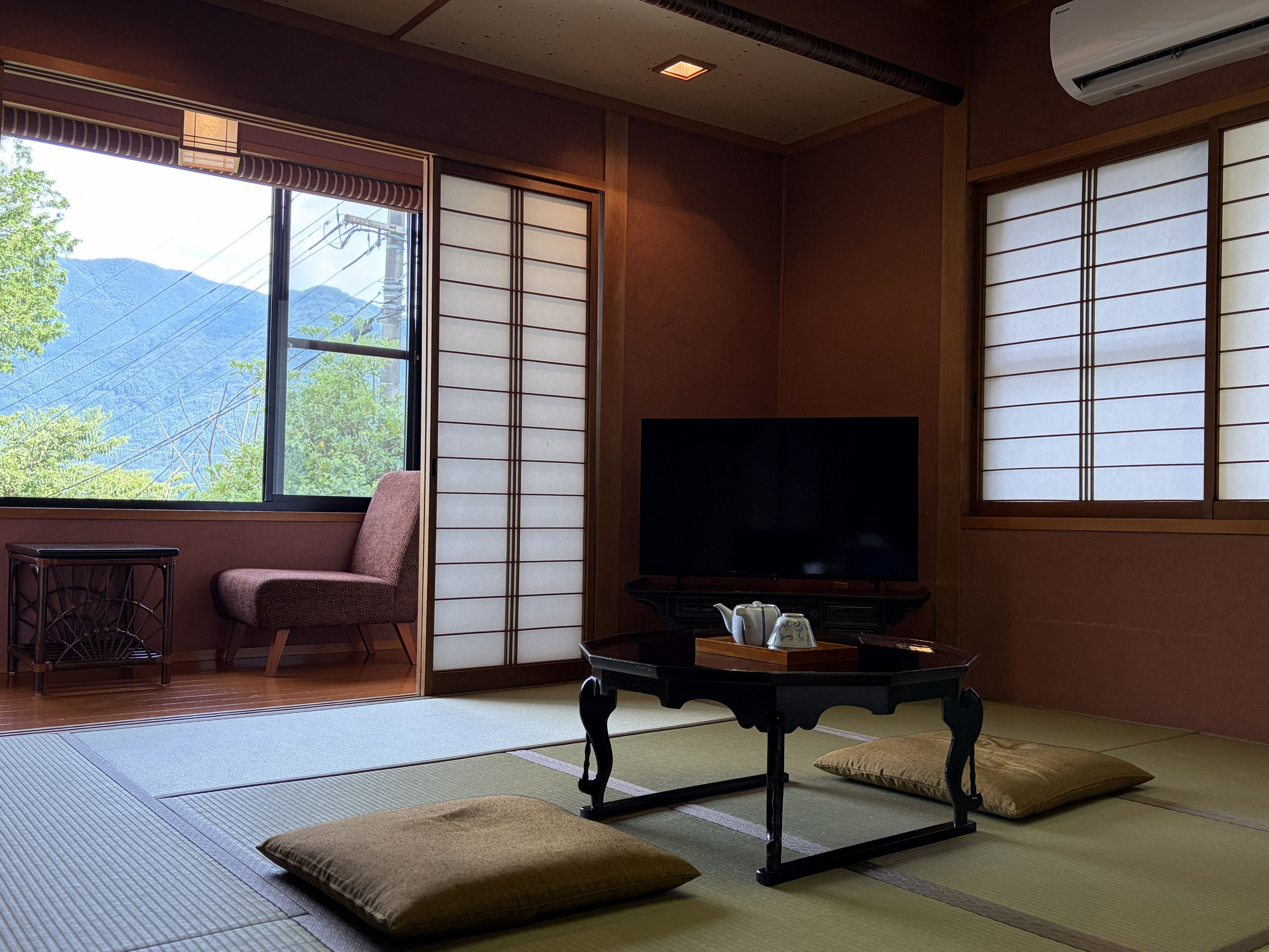 A traditional Japanese room with tatami flooring, a television, a low table with a teapot, and a view of mountains through large windows.