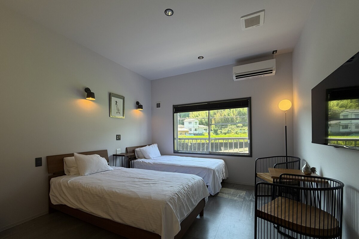 A hotel room with two beds, a desk, a chair, a window with a view, and a mounted TV.