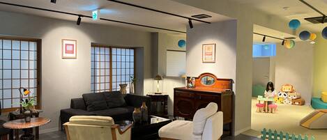 A modern living room with a sofa, armchair, and a small table. There is a wooden cabinet with a mirror and a lamp. The room has large windows with a view of the outside.