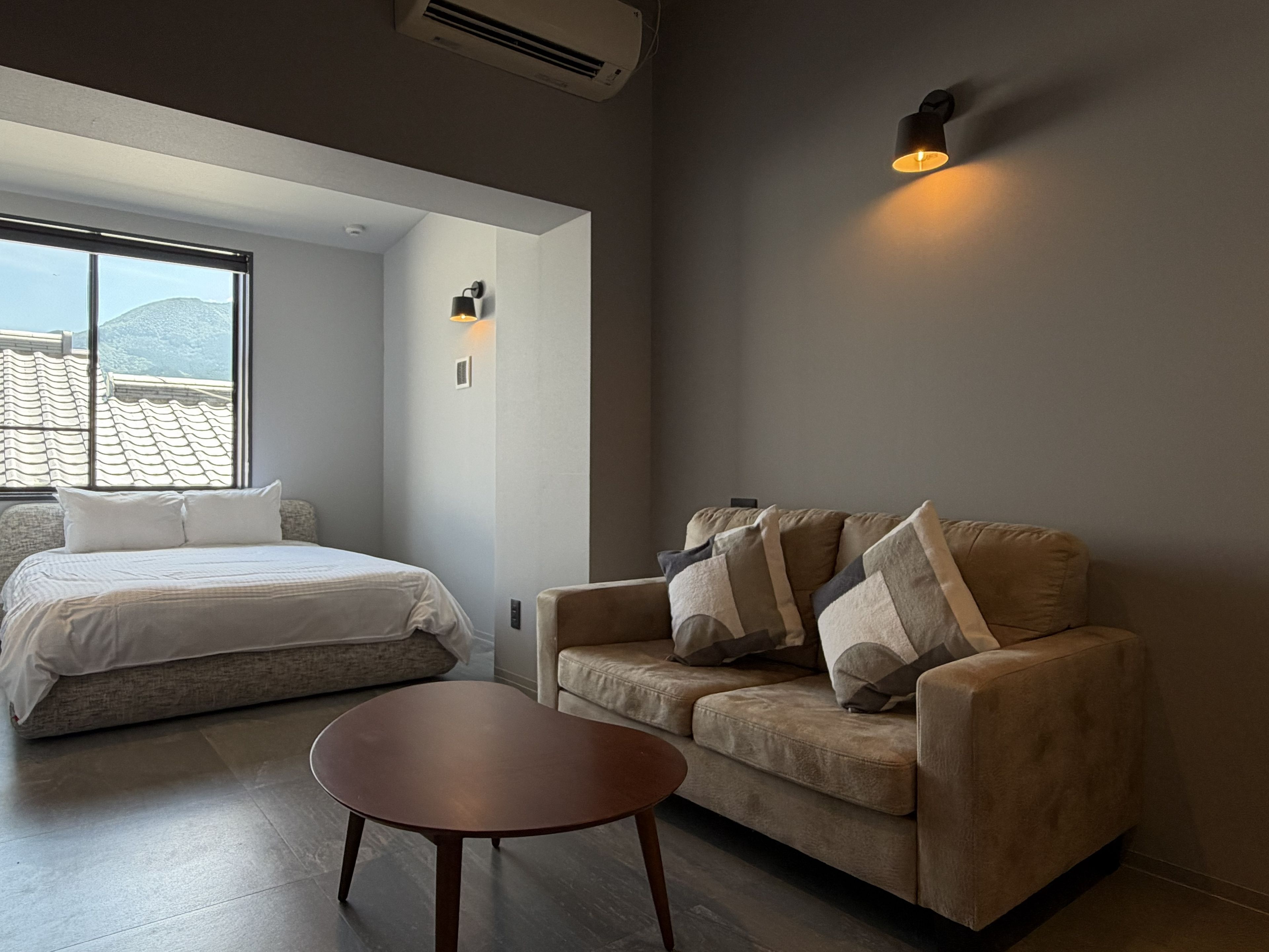 A bedroom with a bed, a sofa, and a small table. There is a window with a view of a building.