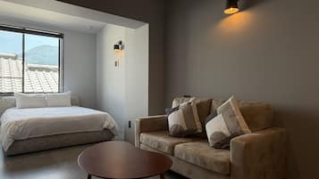 A bedroom with a bed, a sofa, and a small table. There is a window with a view of a building.