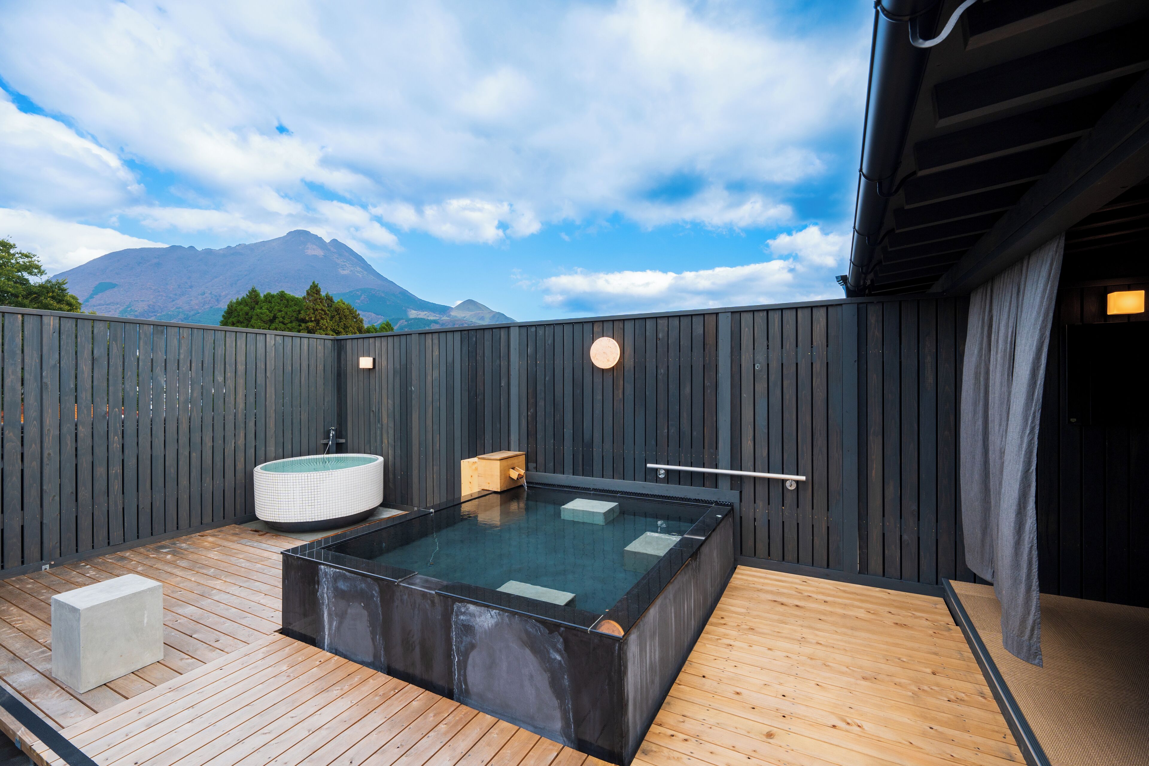 A modern outdoor hot tub area with a wooden deck, a rectangular hot tub, and a cylindrical hot tub, set against a backdrop of mountains and a cloudy sky.