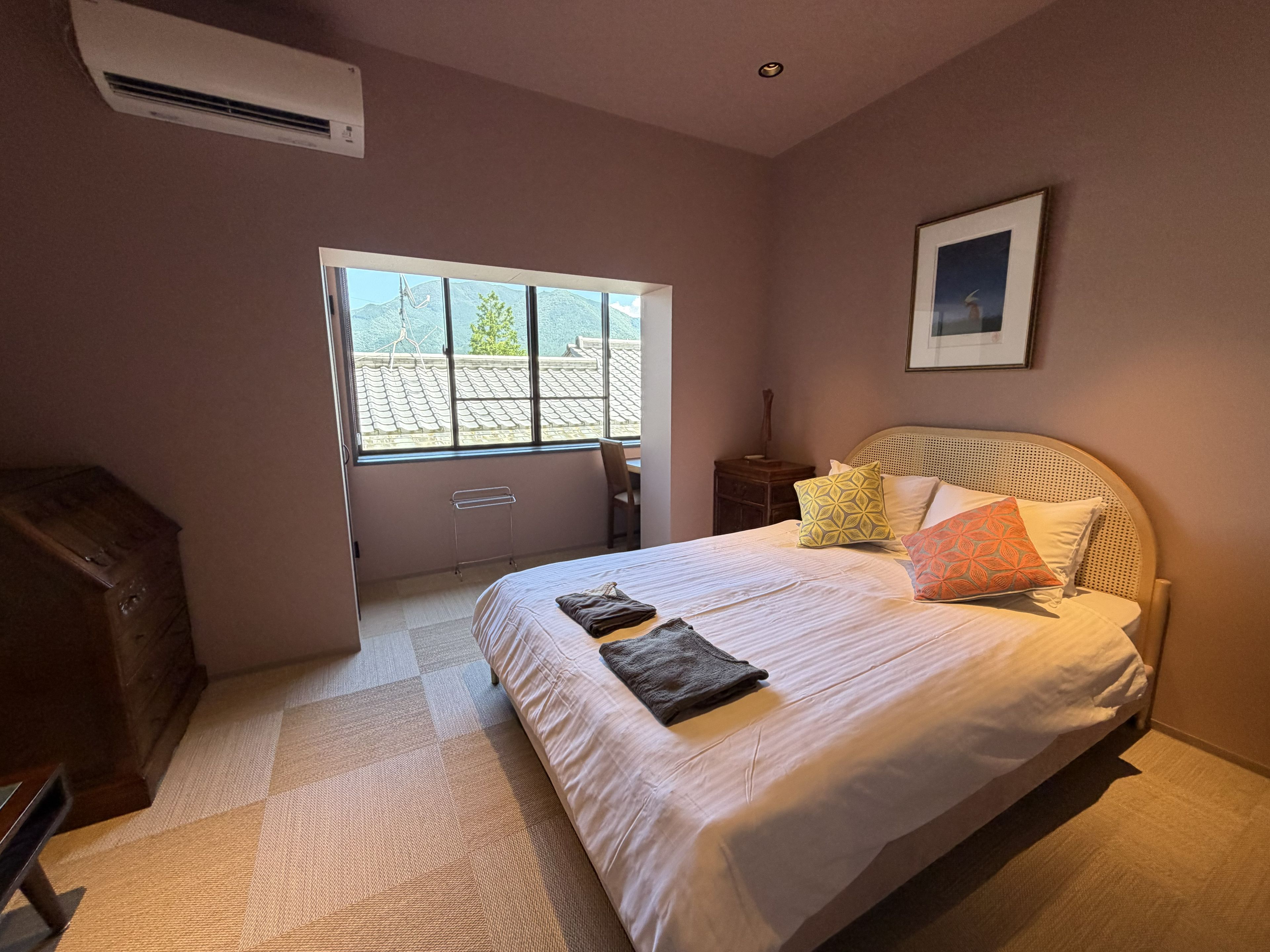 A bedroom with a large bed, a window, a bedside table, and a wall-mounted air conditioner.