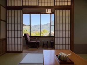 A traditional Japanese room with tatami flooring, a low table, a chair, and a view of a mountainous landscape through large windows. - Ryokan Hachisunoito (Yufu)