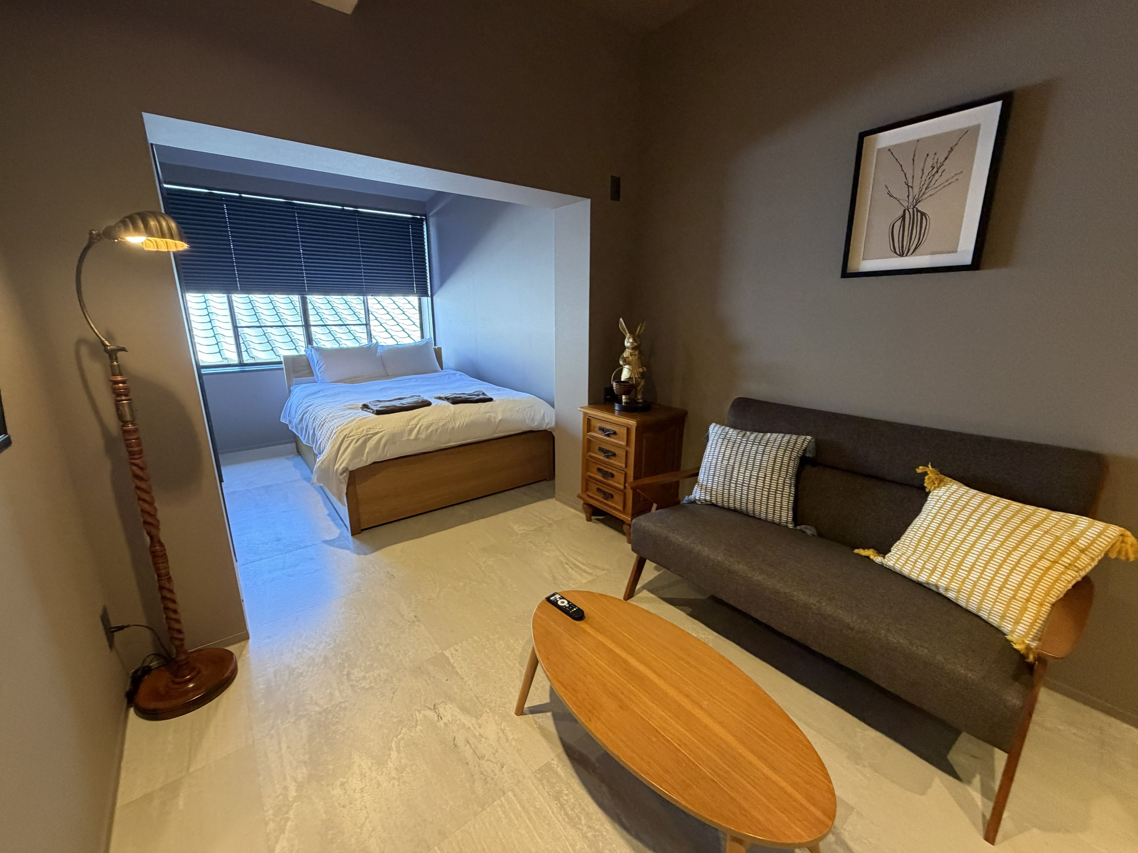 A bedroom with a bed, a sofa, a wooden coffee table, a floor lamp, and a window with blinds.