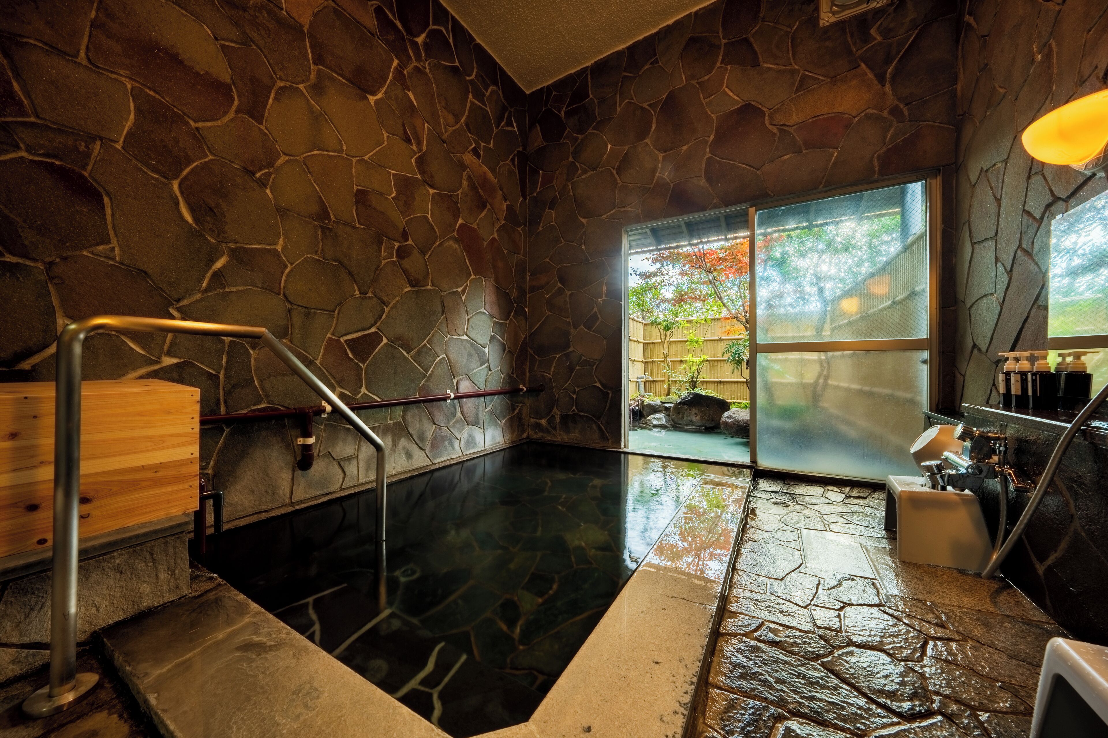 A stone-walled hot spring bath with a wooden bench and a metal railing.