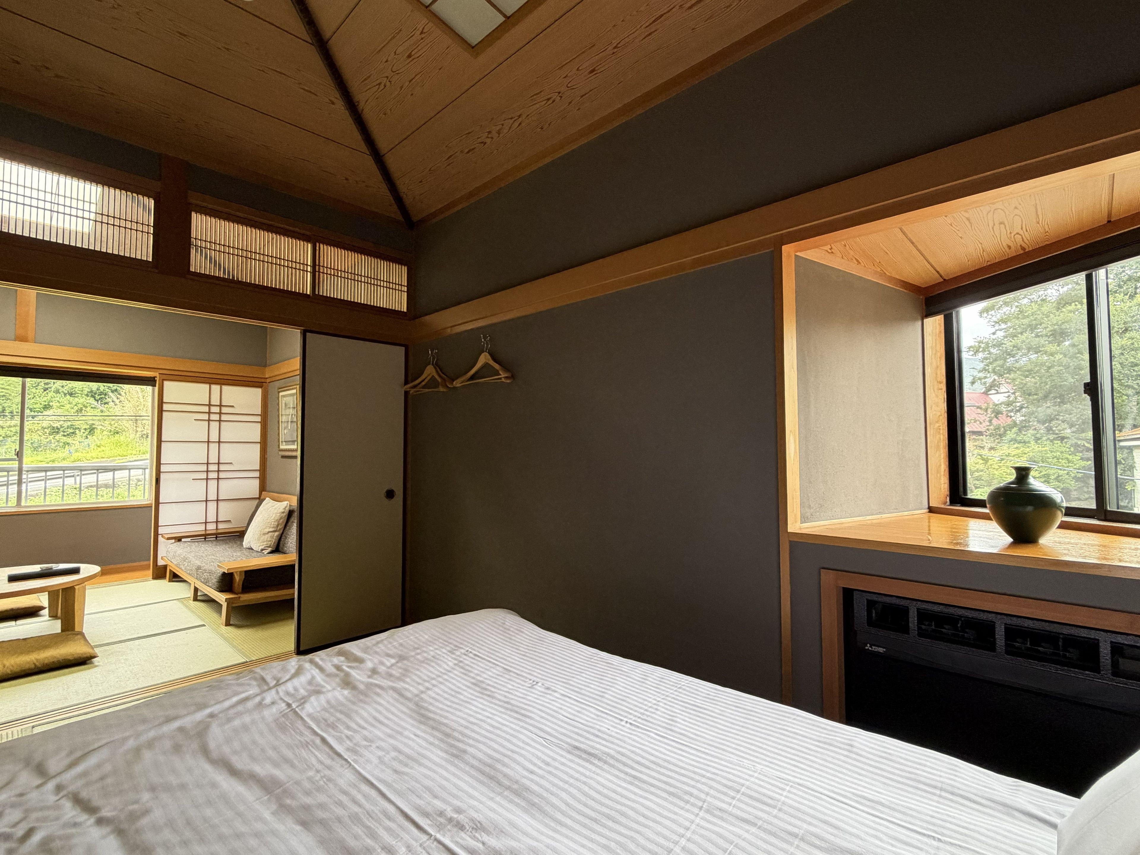 A traditional Japanese-style bedroom with tatami flooring, a low wooden bed, a sliding shoji screen door, and a view of greenery outside.