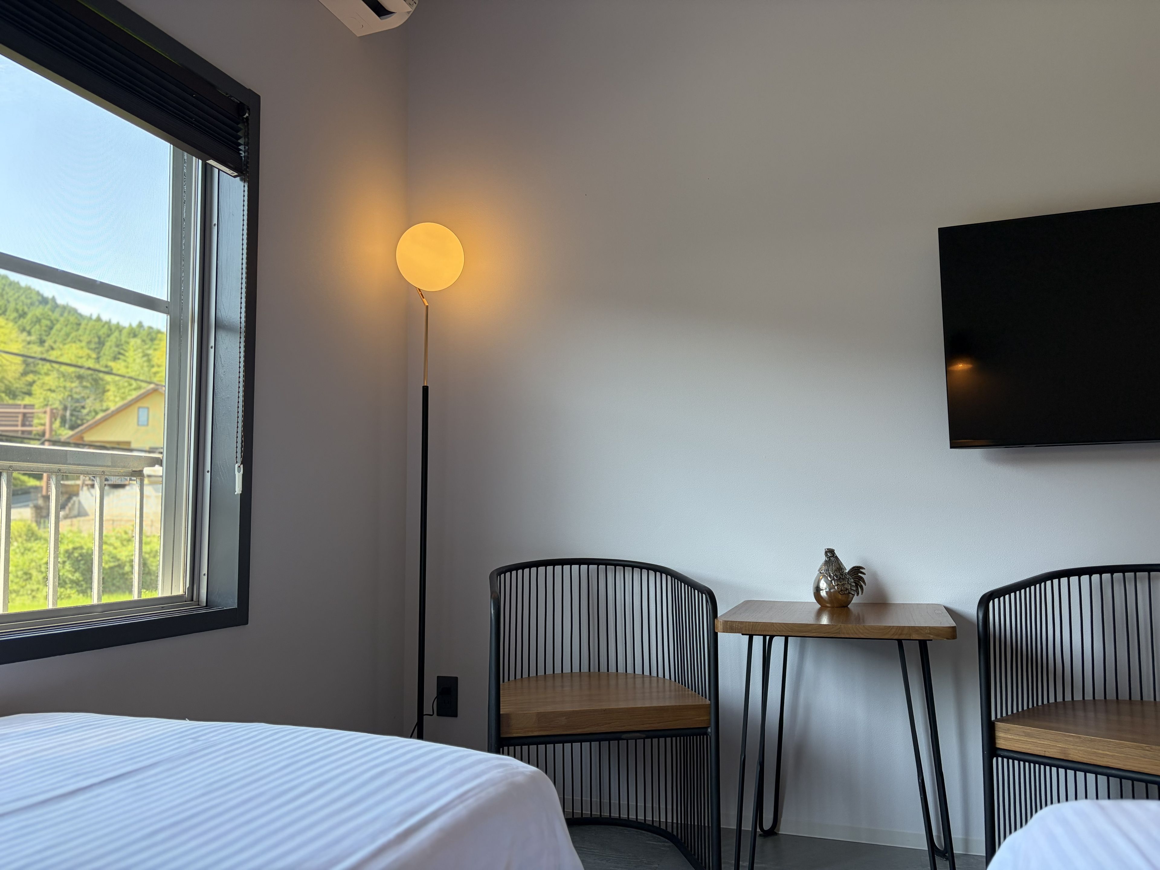 Ein Hotelzimmer mit einem Bett, zwei Stühlen, einem kleinen Tisch, einem am Wand montierten Fernseher und einer Stehlampe.