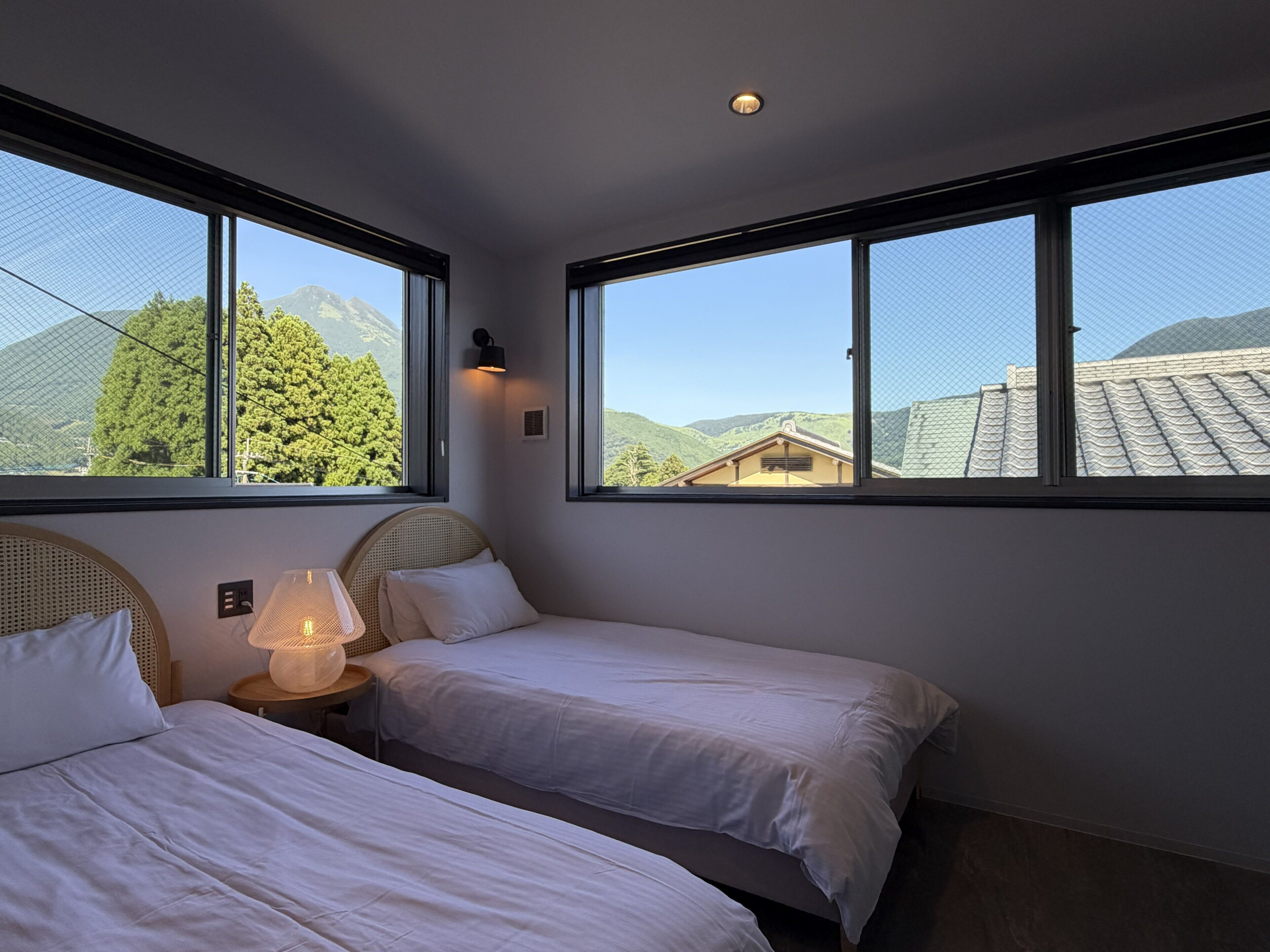 A bedroom with two beds, a nightstand with a lamp, and large windows offering a mountain view.