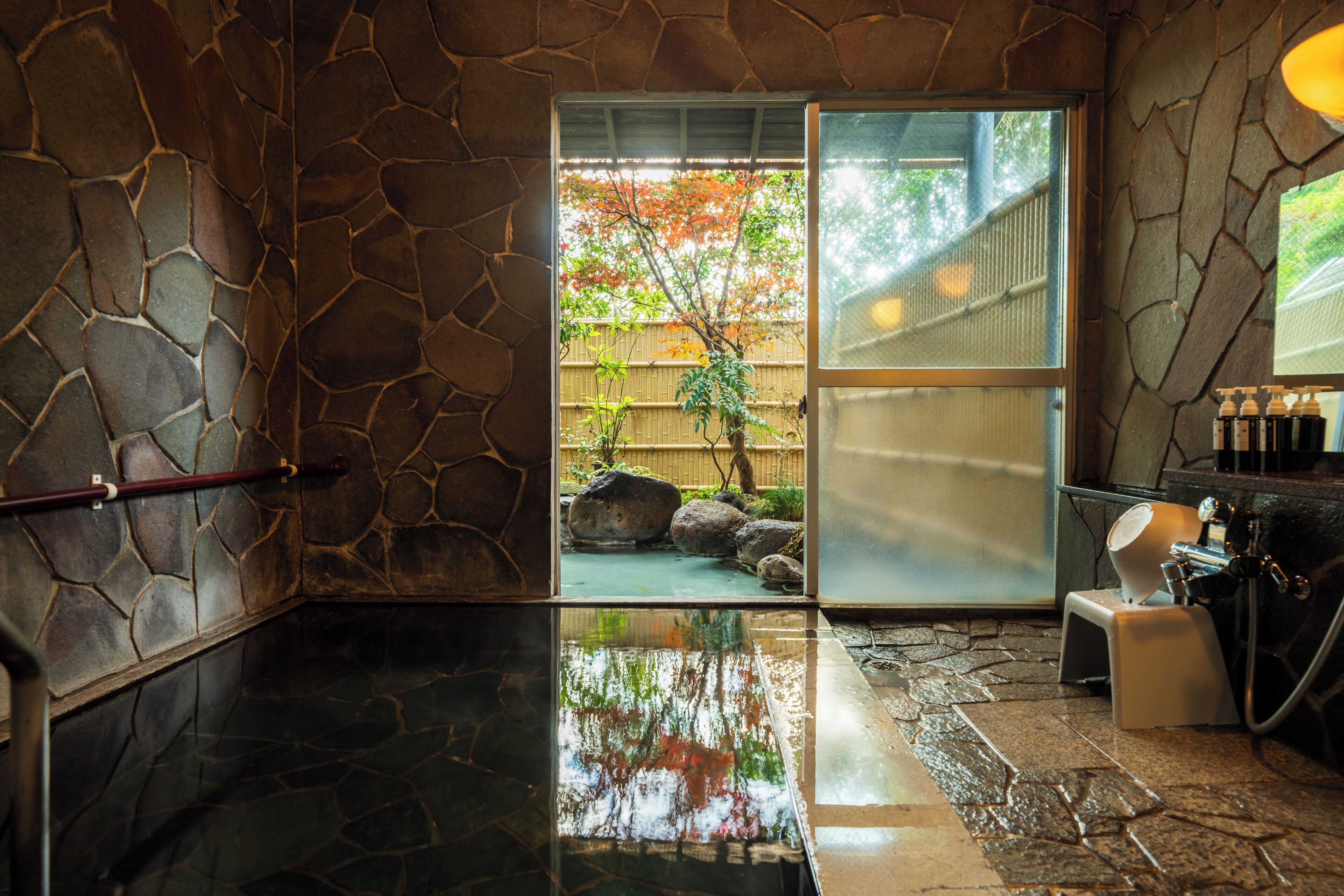 A stone-walled room with a glass door leading outside, a small stool, and a water feature.