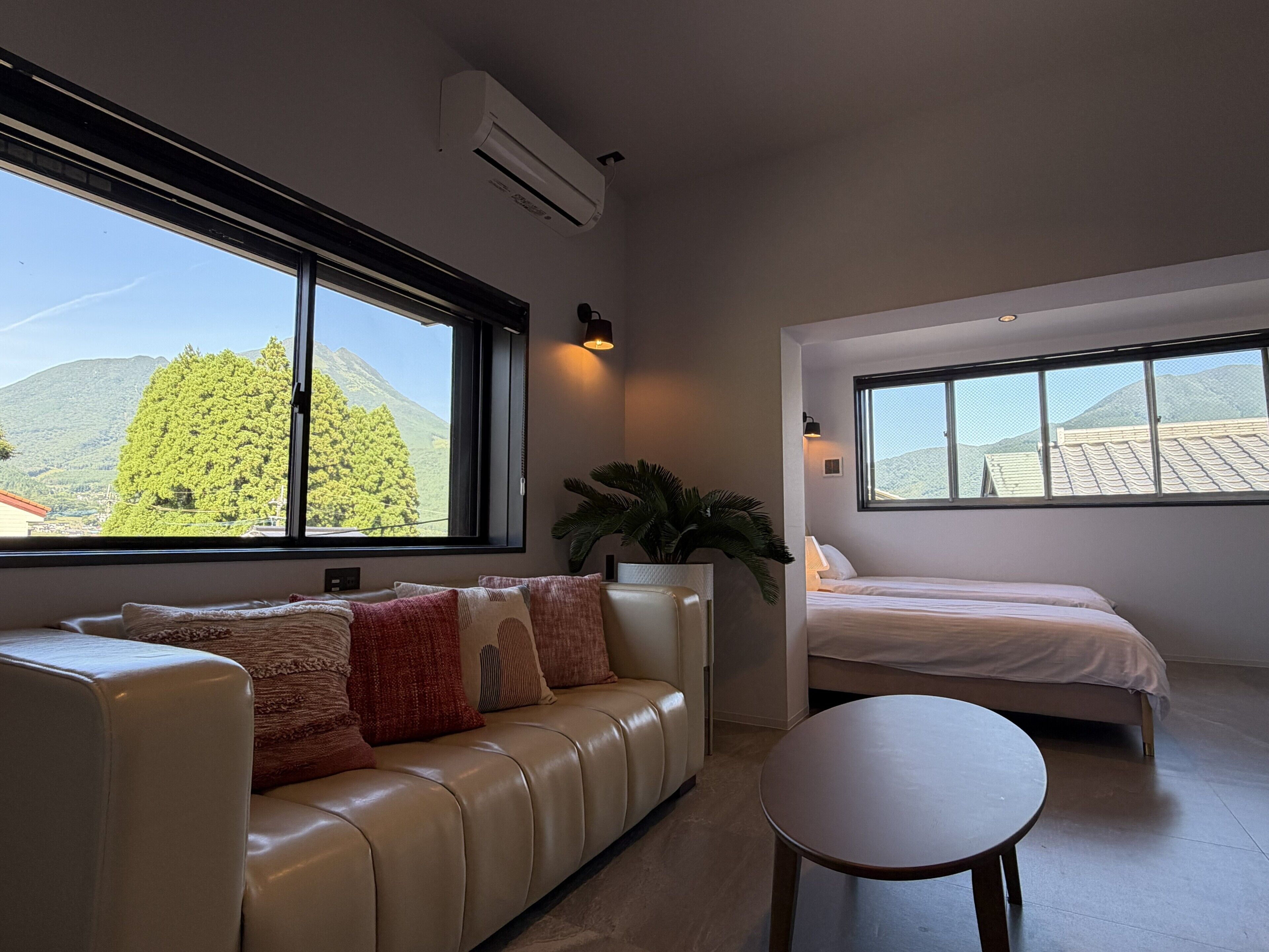 A modern room with a sofa, a round table, and a bed, featuring large windows with a mountain view.