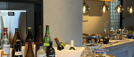 A bar area with various bottles and glasses, a refrigerator, and a shelf with decorative items.