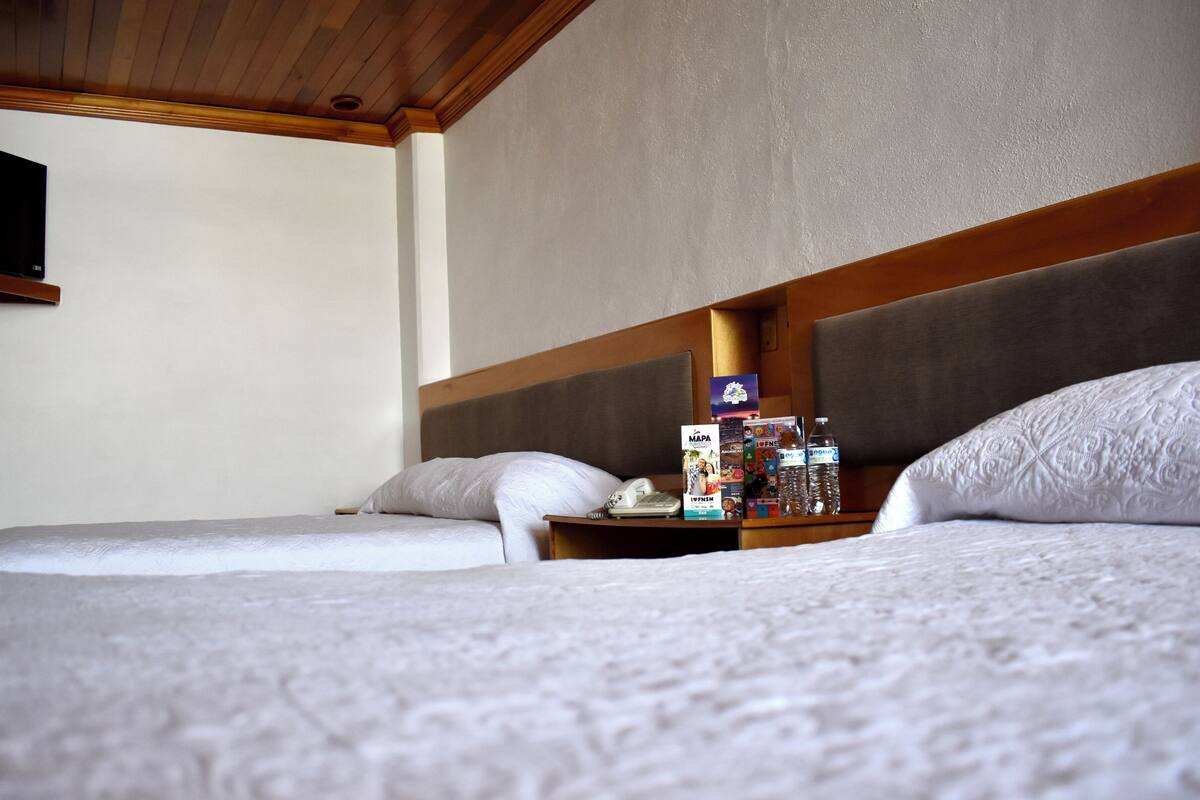 Habitación de hotel con dos camas, un cabecero de madera, una mesita de noche con teléfono y revistas, y un televisor montado en la pared.