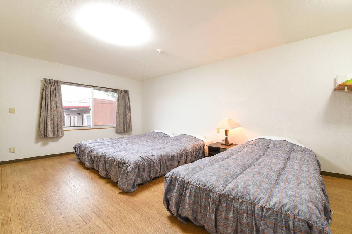 Una habitación con dos camas, una ventana con cortinas, una mesita de noche con lámpara y un estante con objetos.