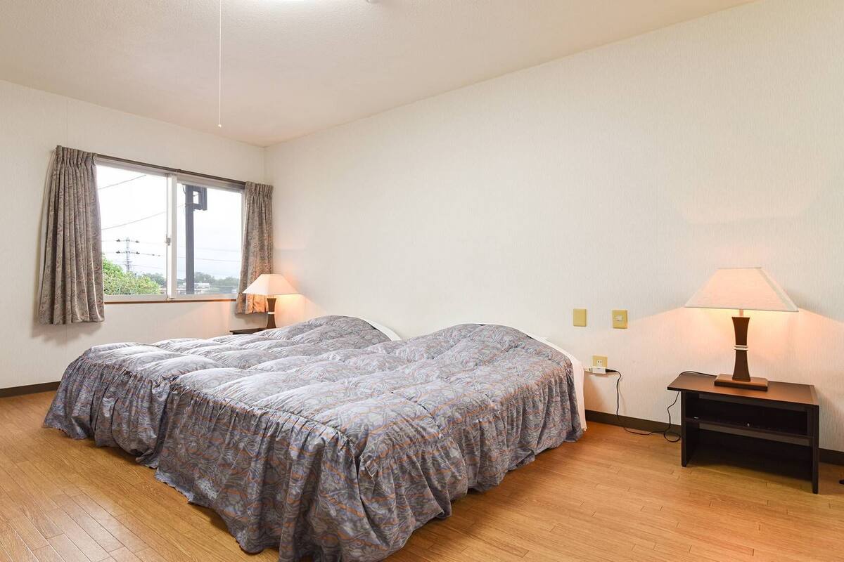 Una habitación con dos camas, una ventana con cortinas, una mesita de noche con lámpara y suelo de madera.